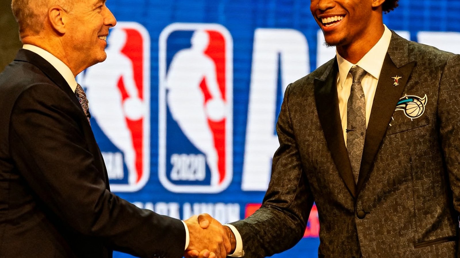 2025年NBA选秀大会现场，新秀球员与总裁握手合影，身穿球队帽子面带笑容